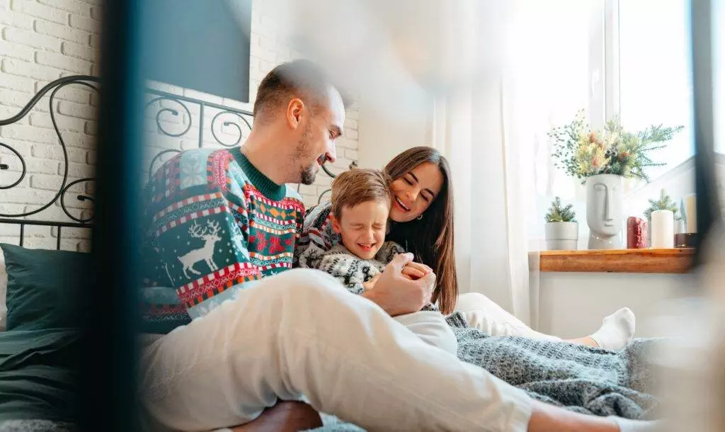 Family laughing together on a bed, enjoying a cozy moment by a window.